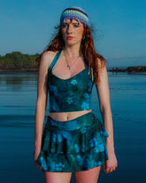 Woman in a teal outfit with a crochet cap standing by a body of water.