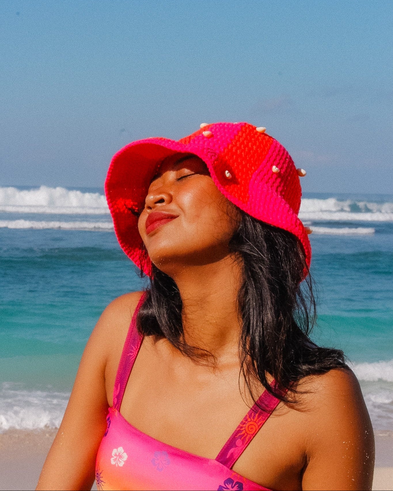 Woman wearing a red hat and pink top on a beach with blue sky and ocean.
