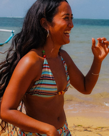 A woman smiling on a beach wearing a colorful striped bikini top and matching bottoms, with ocean water and sand in the background.