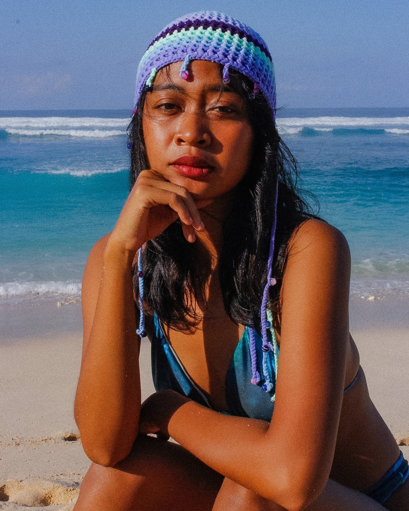 Woman sitting on a beach wearing a blue and purple knit hat with braided ends.