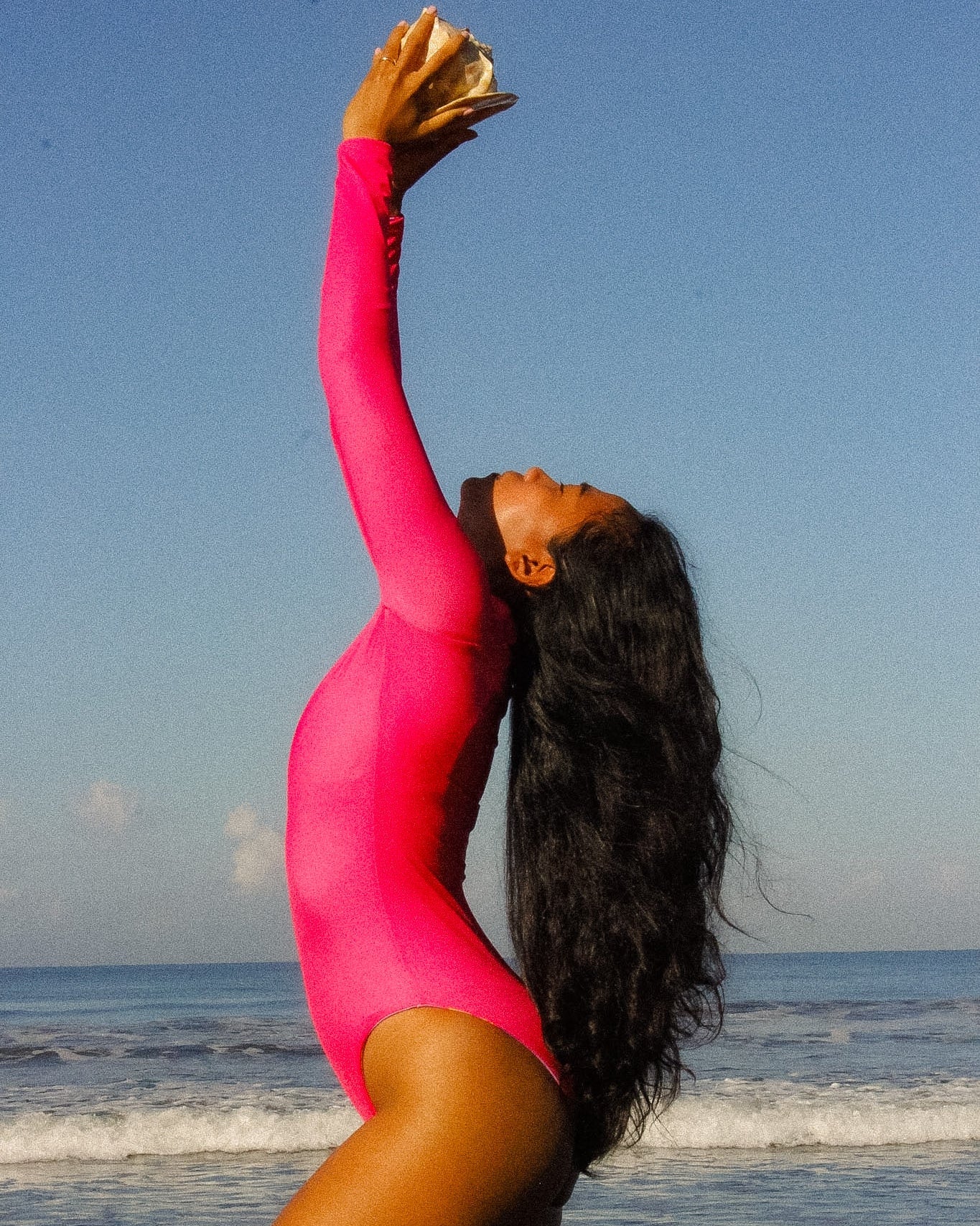 Person in a bright pink swimsuit holding a shell above their head on a beach.