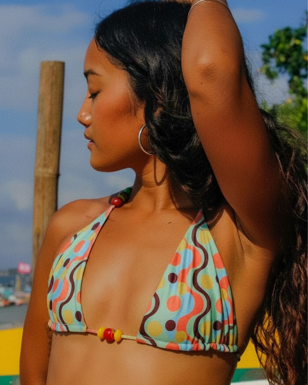 A woman wearing a colorful triangle bikini top with a retro wavy pattern in orange, yellow, blue, and brown on a light blue background, photographed outdoors with a wooden post and sky behind her.